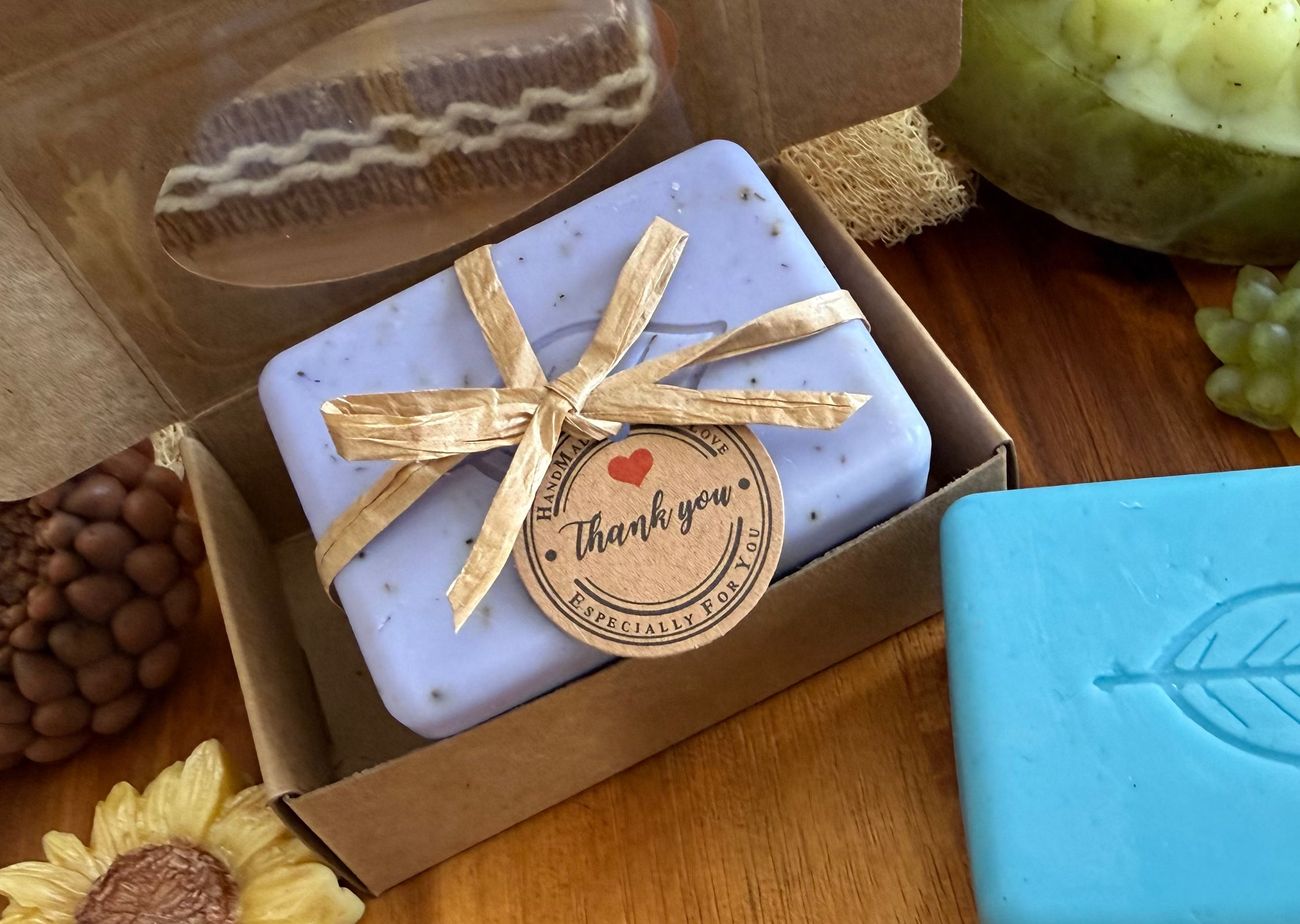 A gift box of artisanal soaps displayed on a wooden table. The box is open to reveal a lavender soap in a clear window and a lavender soap tied with a twine bow and a 'Thank you' tag.